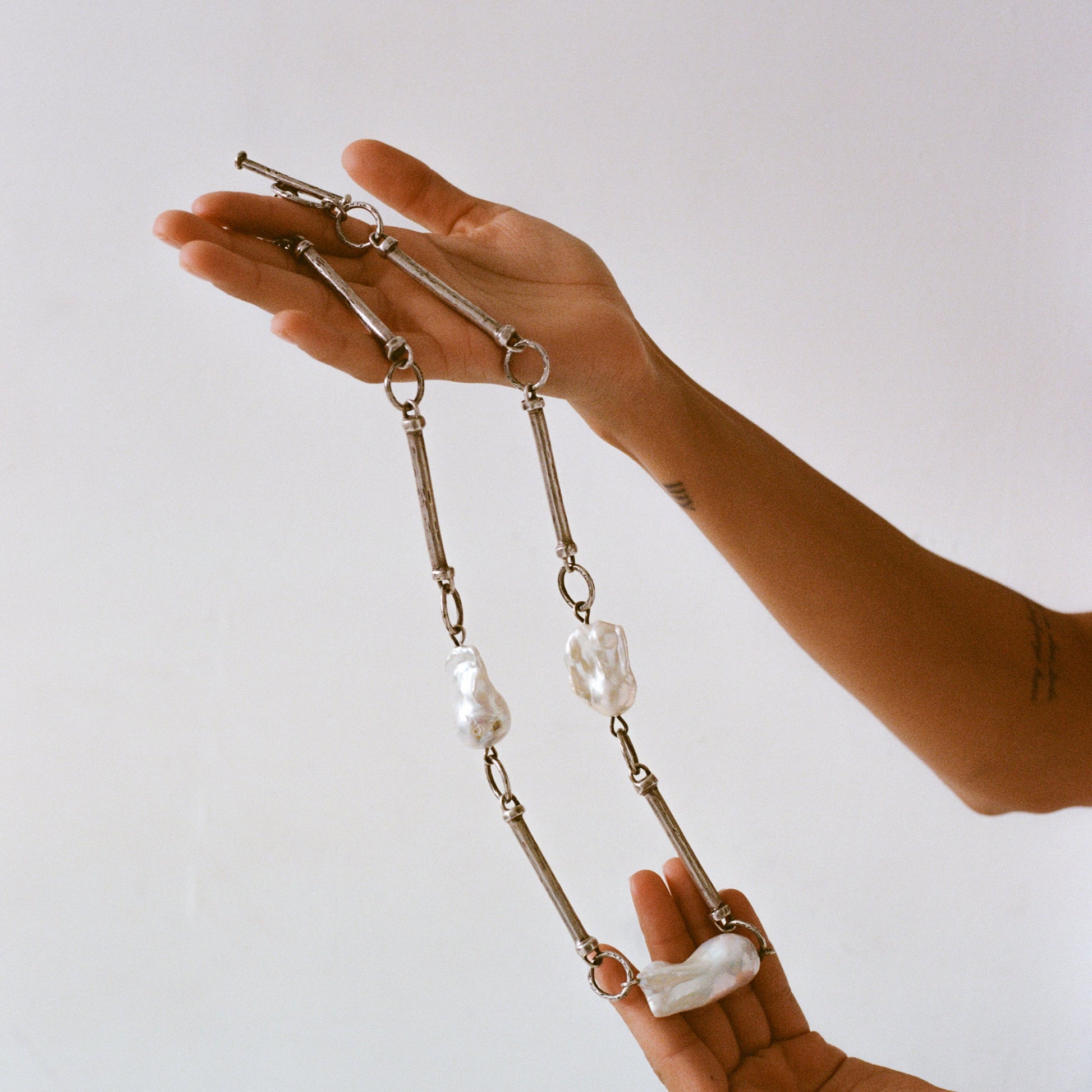 A close-up image of a sterling silver and baroque pearl necklace held in a person's hand, featuring a series of linked silver bars with baroque pearl accents.