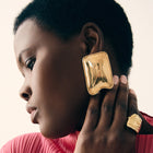 Woman wearing large gold earrings and a matching ring, with a pink top.